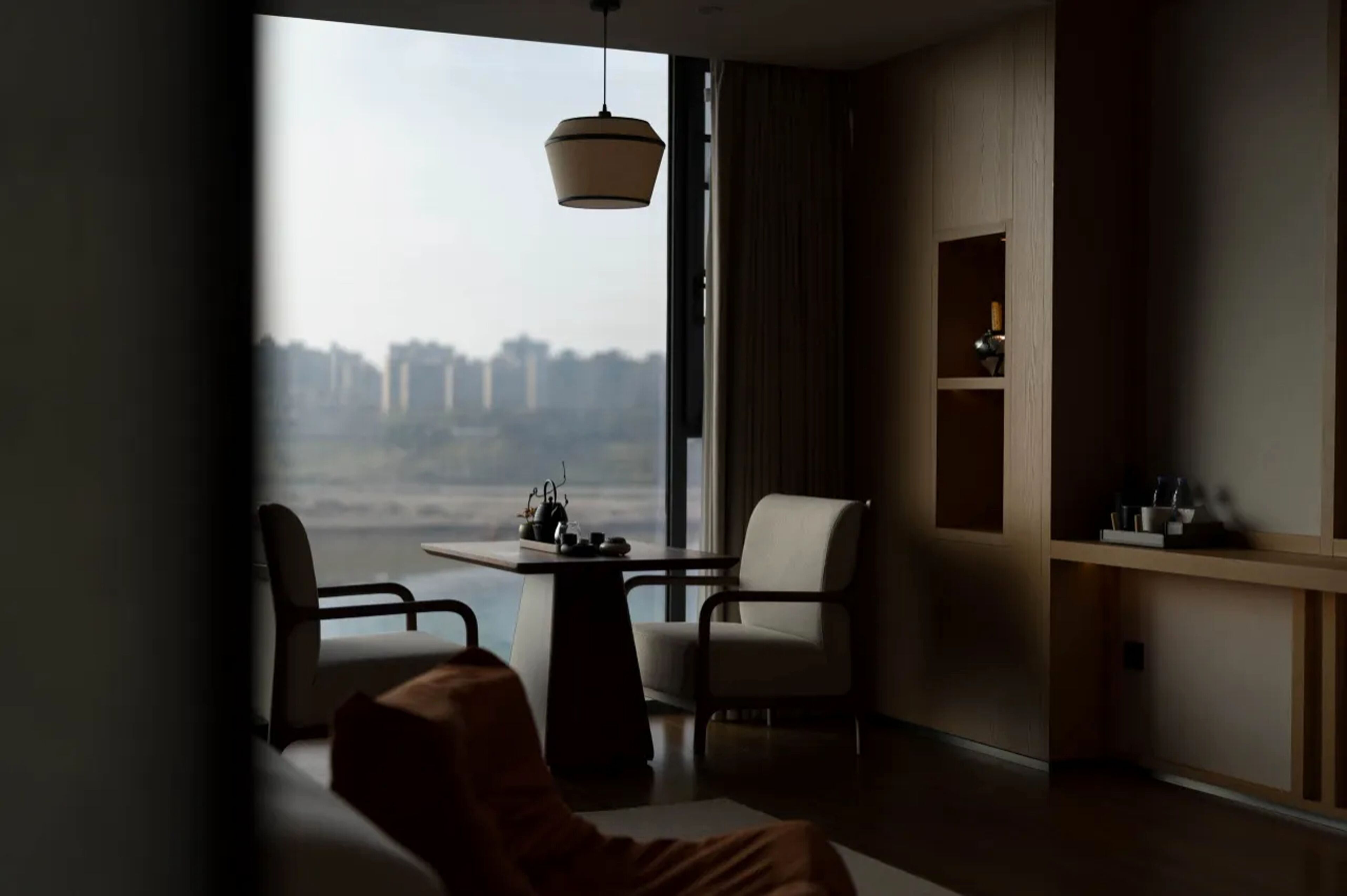 A modern hotel room with a large window, a dining table, two chairs, a sofa, a hanging light fixture, and built-in shelving.