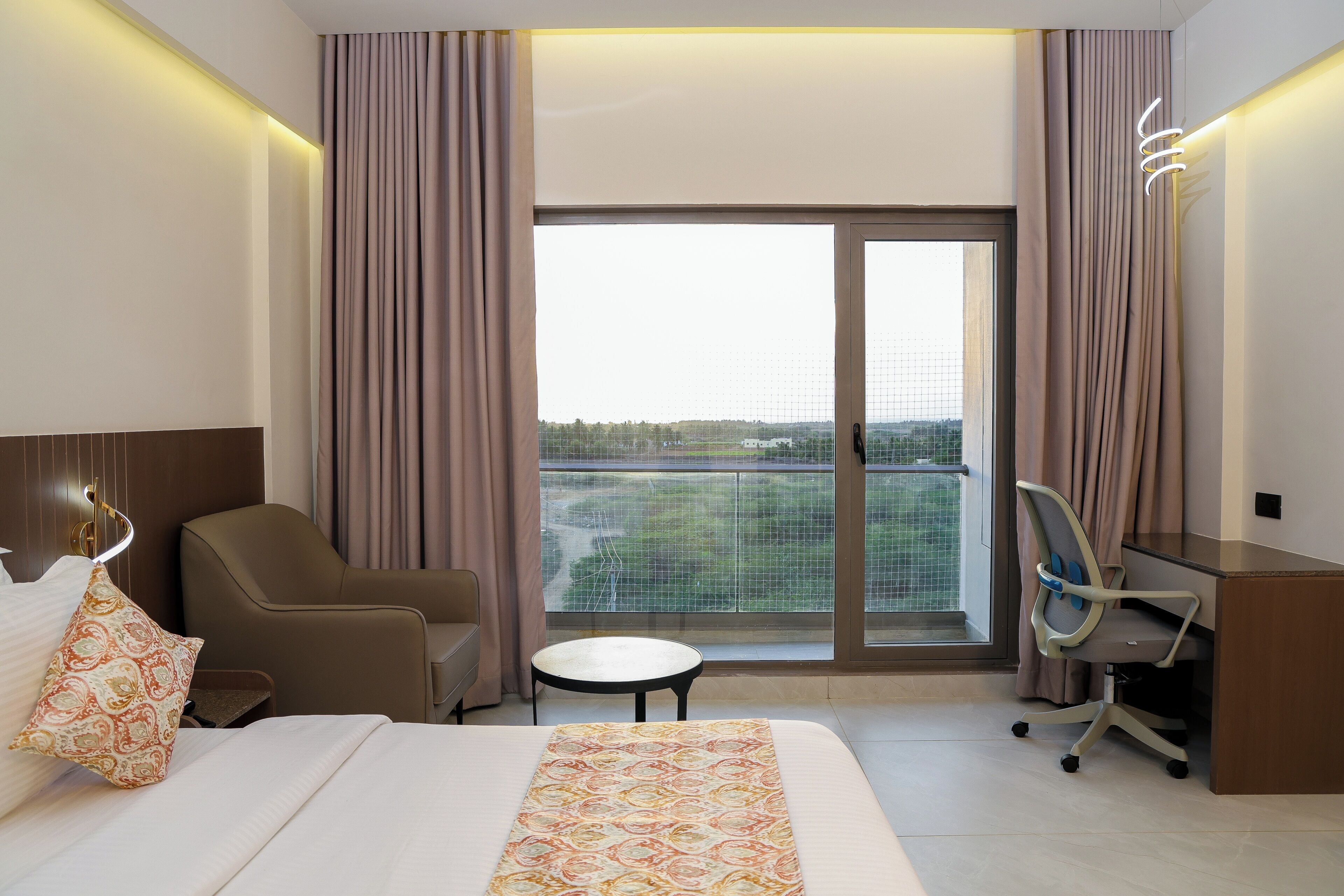 Habitación de hotel con cama, escritorio, silla, una mesita y vistas a través de un ventanal amplio.