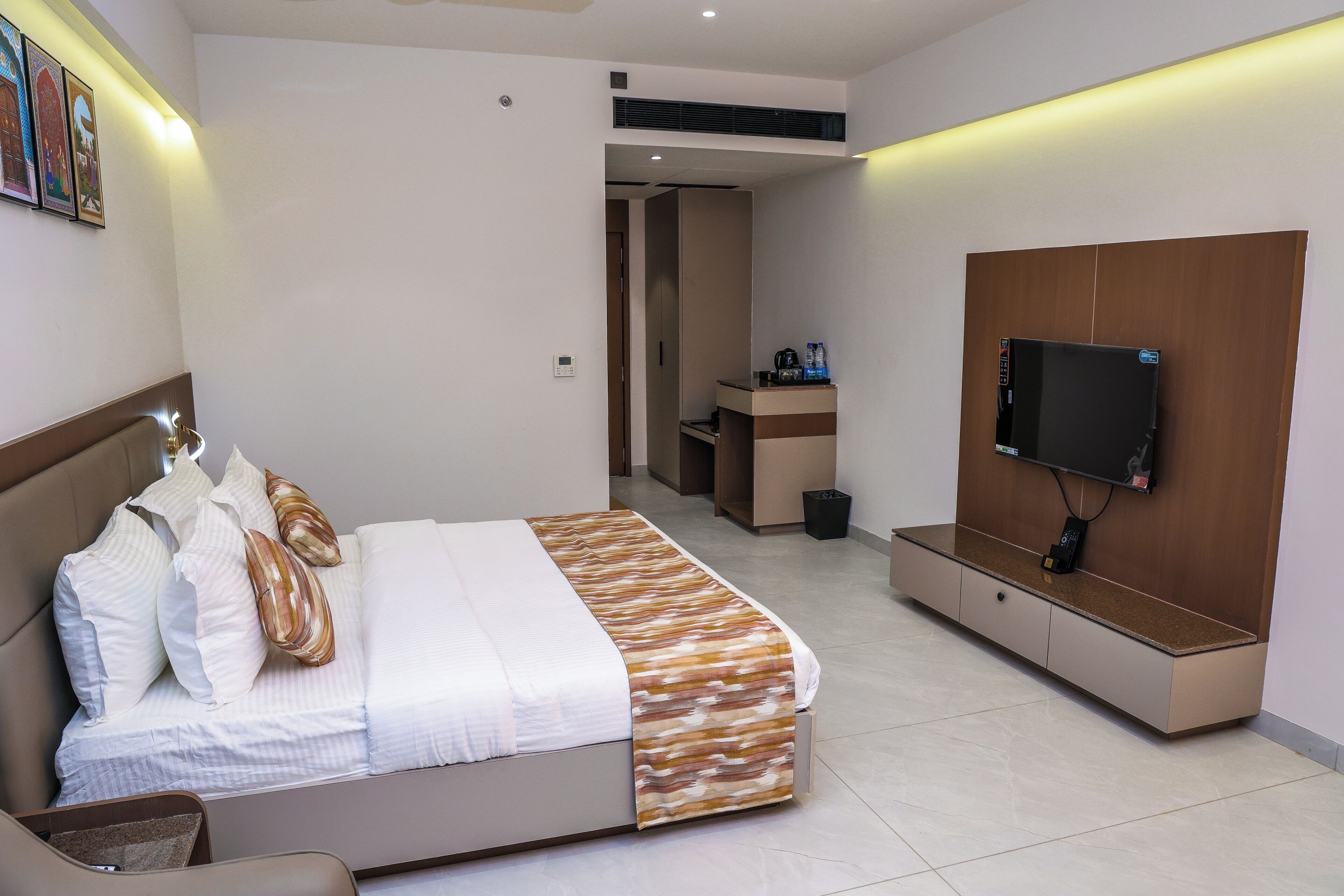 A modern hotel room with a bed, a TV mounted on a wooden panel, a small desk, and a bathroom visible through an open door.