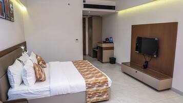 A modern hotel room with a bed, a TV mounted on a wooden panel, a small desk, and a bathroom visible through an open door.