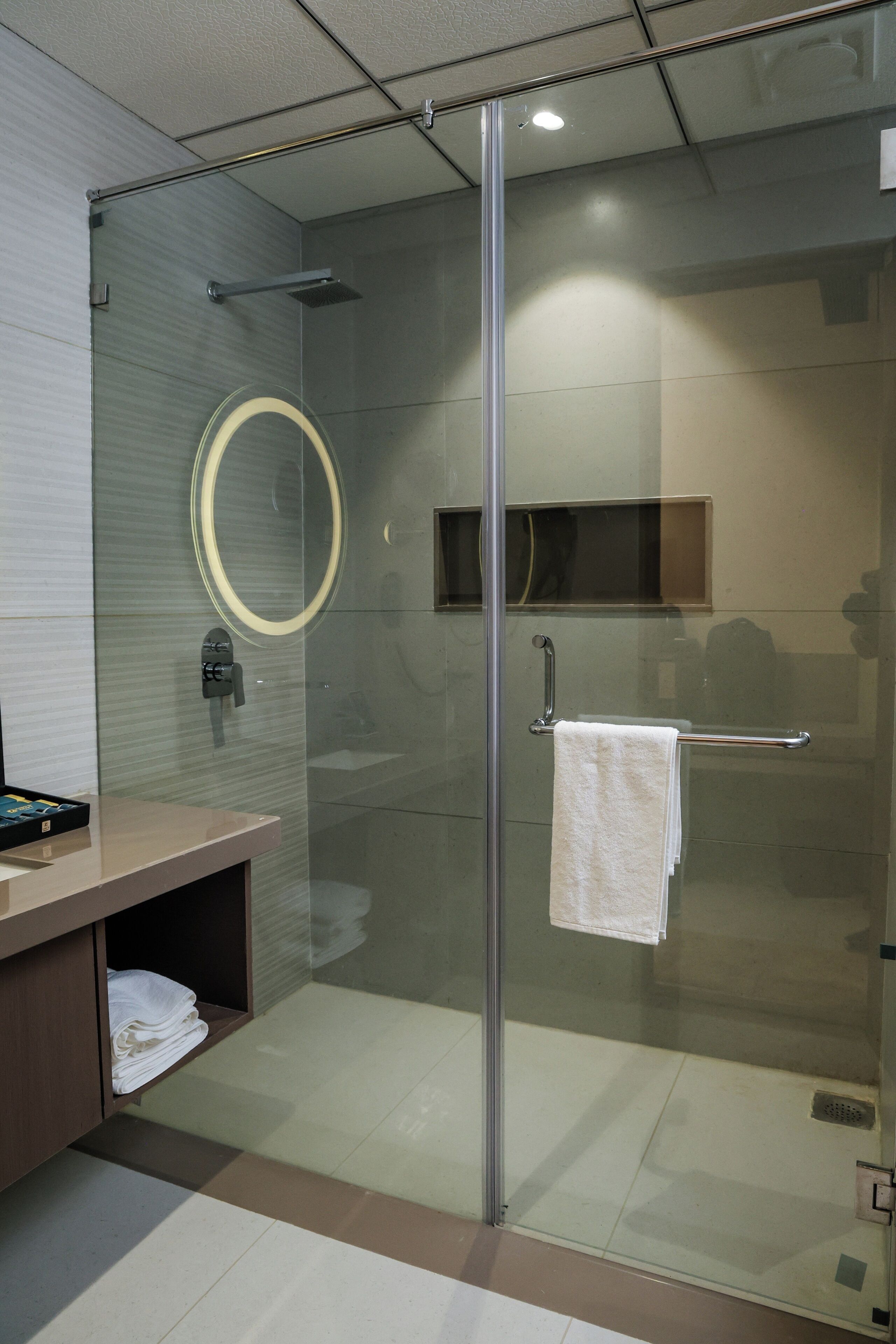 A modern bathroom with a glass shower enclosure, a round mirror, a towel rack, and a wooden vanity with a towel shelf.