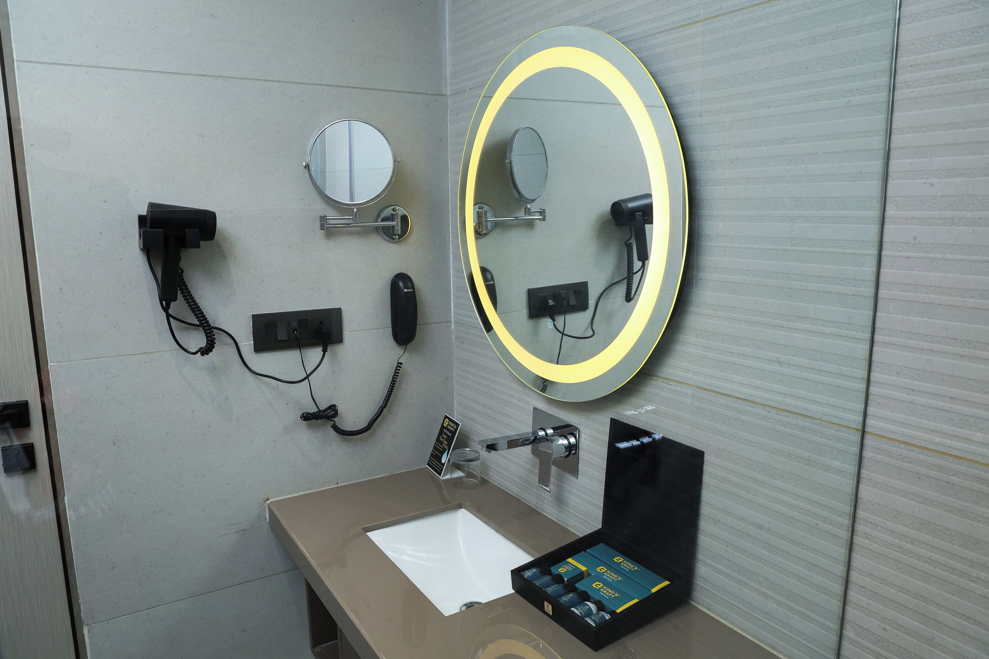 A bathroom with a sink, two mirrors, a hairdryer, a telephone, and a soap dispenser.