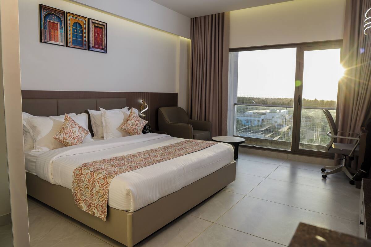 Une chambre d’hôtel avec un grand lit, une chaise, une petite table et une vue sur la ville grâce à de grandes fenêtres.