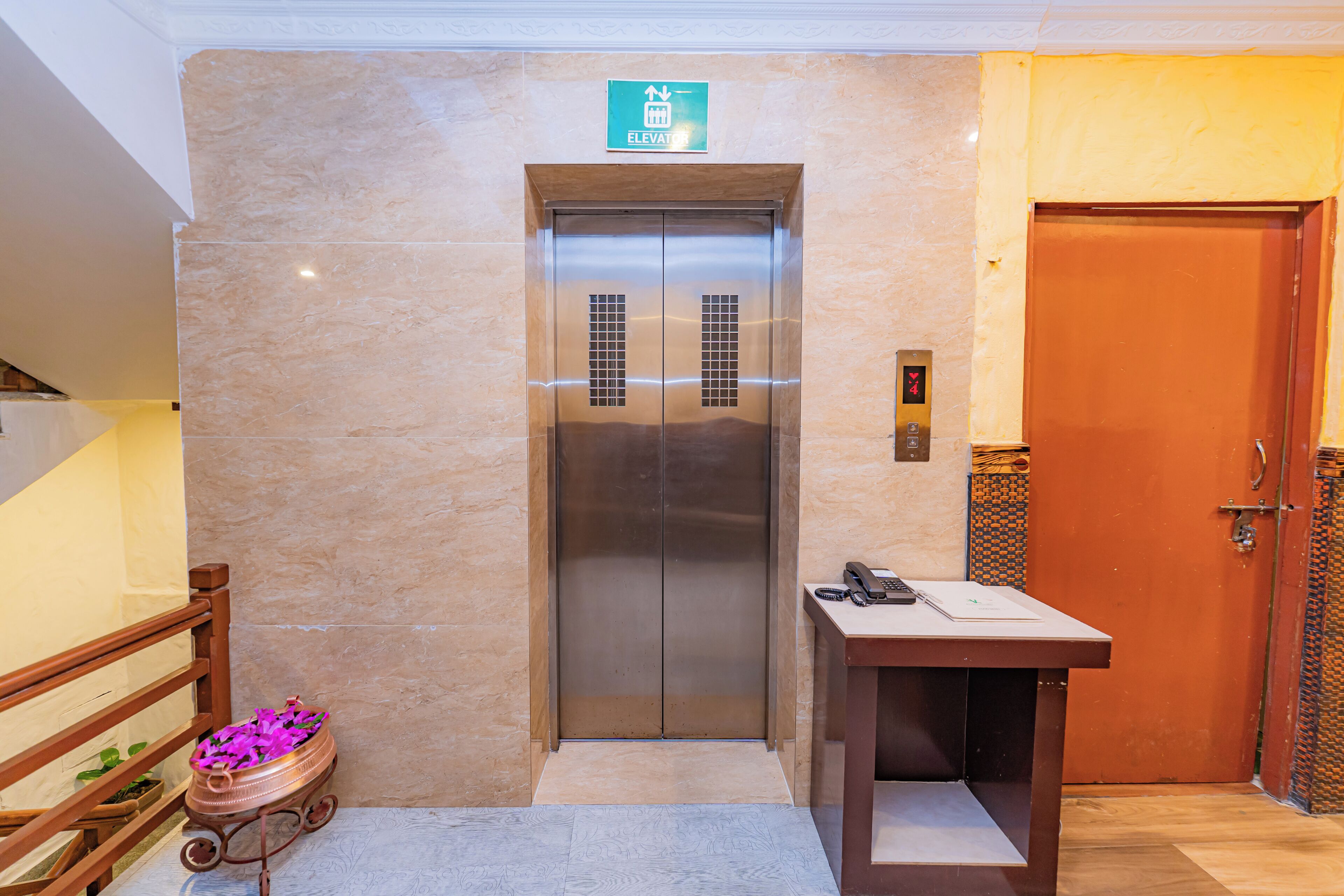 An elevator with a green emergency exit sign, a wooden door to the right, and a small table with a phone and papers.