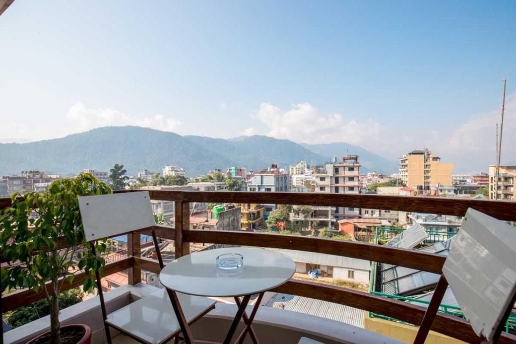 A balcony with a table and chairs, overlooking a cityscape with buildings and mountains.
