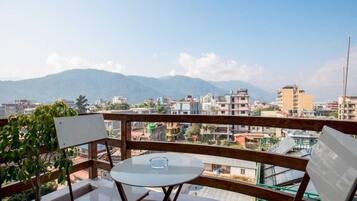 A balcony with a table and chairs, overlooking a cityscape with buildings and mountains.