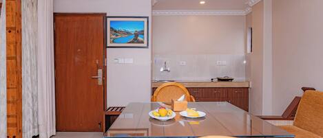 A dining area with a glass table, wooden chairs, a wooden door, and a kitchen in the background.