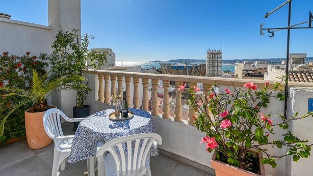 Ein Balkon mit Tisch und Stühlen, Blumentöpfen und Blick auf das Meer und die Gebäude.