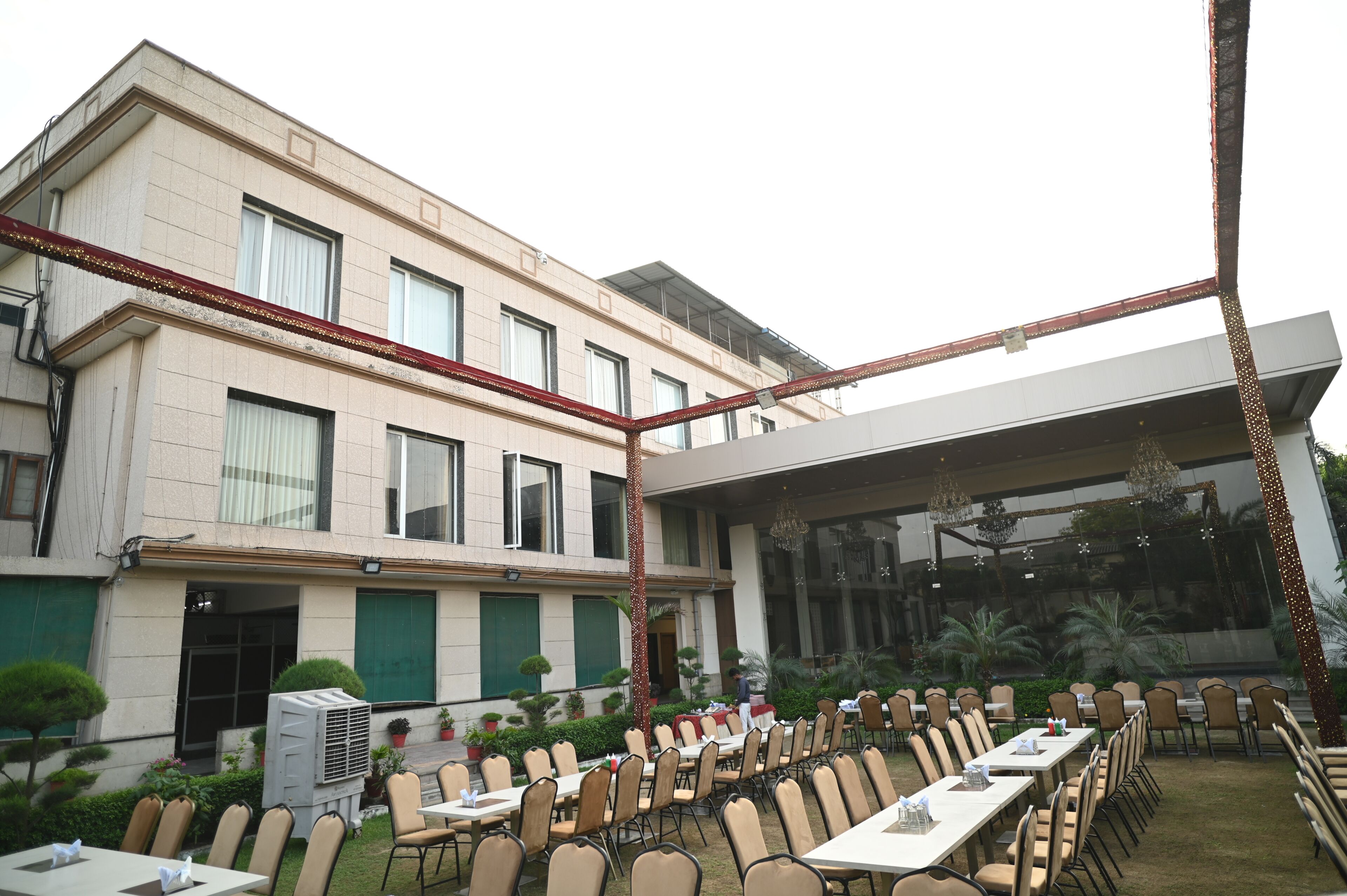 Un hotel con zona de comedor al aire libre, varias mesas y sillas, y un edificio grande con ventanas y una sección cubierta.