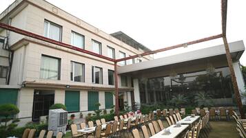 Un hotel con zona de comedor al aire libre, varias mesas y sillas, y un edificio grande con ventanas y una sección cubierta.