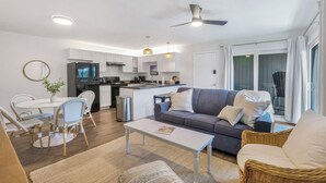 Ein modernes Wohnzimmer mit Sofa, Couchtisch, Esstisch und Stühlen. Es gibt eine Küchenzeile mit Schränken und Geräten.