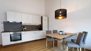 A modern kitchen with white cabinetry, a built-in oven, and a dining area with a wooden table and grey chairs. - Apartment in Milders With Stubai Supercard (Neustift Im Stubaital)
