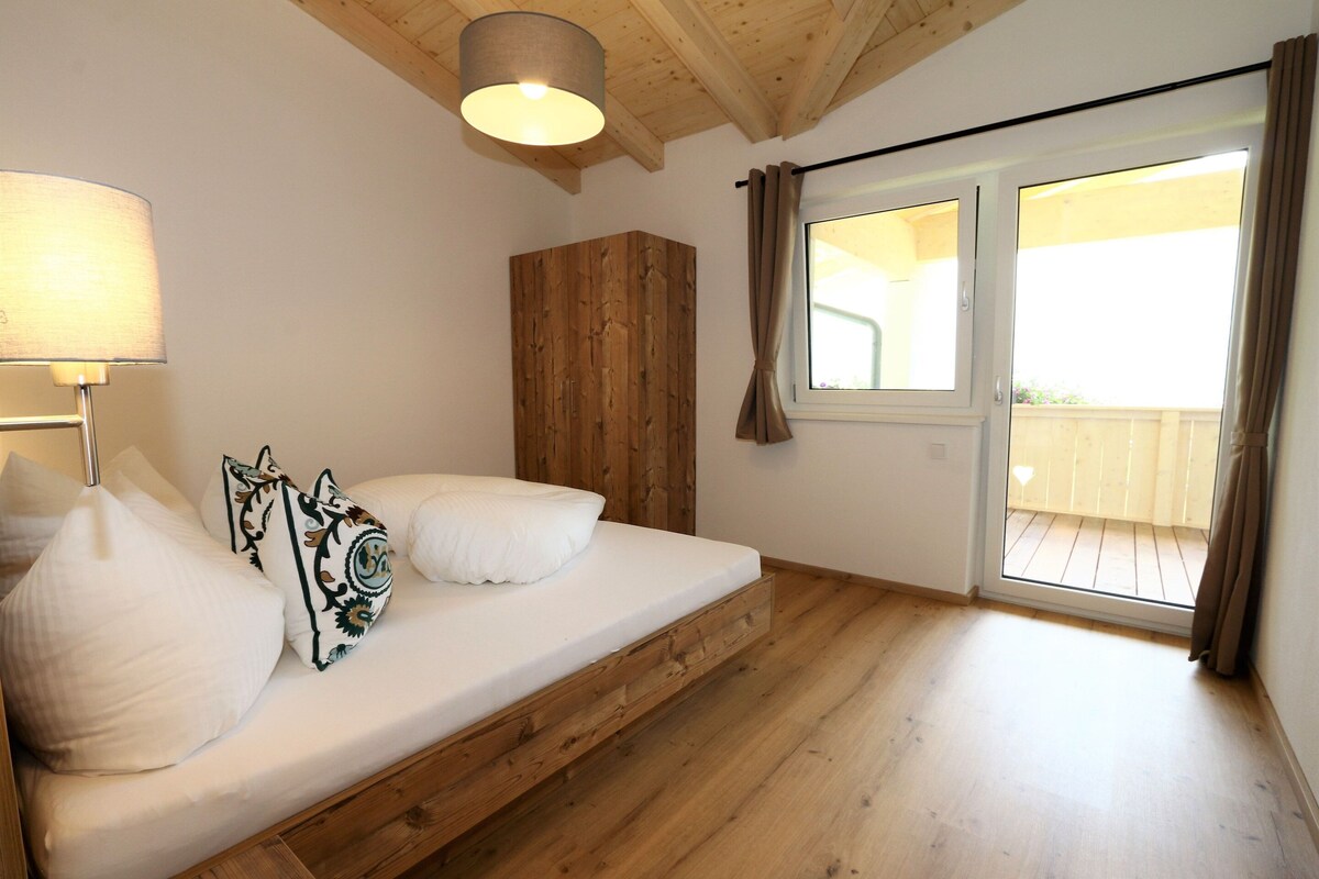 Ein Schlafzimmer mit einem hölzernen Bett, einem Nachttisch, einer Lampe und einem großen Fenster mit Vorhängen.