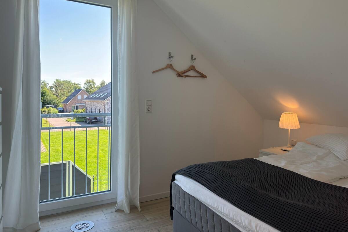 Un dormitorio con cama, una lámpara colgante, ventana con cortinas y vista a una casa y jardín.