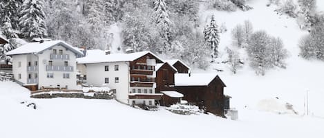 Un pueblo en la montaña cubierto de nieve, con edificios de varios pisos, incluyendo un hotel con balcones y una cabaña de madera.