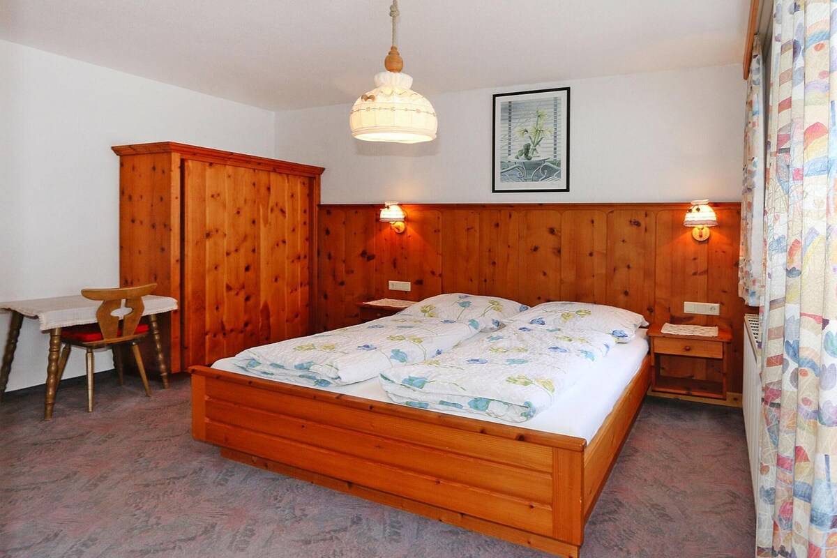 Una cama de madera con colcha estampada, un armario de madera, una mesita con una silla y una lámpara fijada a la pared.