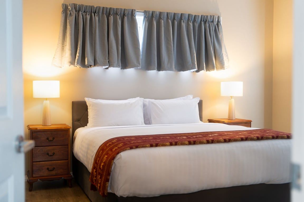 A hotel room with a bed, two bedside tables, a nightstand, a window with curtains, and two wall-mounted lamps.