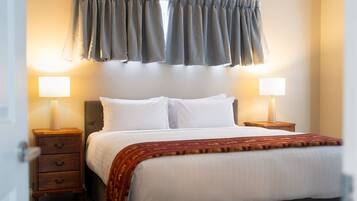 A hotel room with a bed, two bedside tables, a nightstand, a window with curtains, and two wall-mounted lamps.