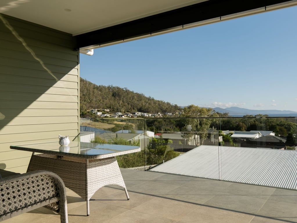 A balcony with a glass table and wicker furniture, overlooking a residential area with houses and a hilly landscape.