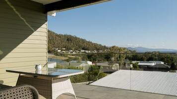 Un balcón con una mesa de vidrio y mobiliario de mimbre, con vistas a una zona residencial con casas y un paisaje montañoso.