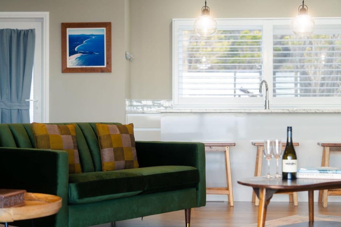 A green sofa with checkered cushions, a wooden table with a bottle and glasses, and a window with blinds.