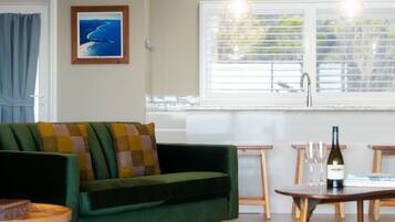 A green sofa with checkered cushions, a wooden table with a bottle and glasses, and a window with blinds.