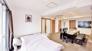 Quarto de hotel com cama, mesa de jantar e cadeiras, televisão e relógio na parede.