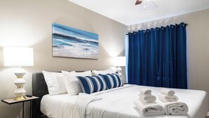 A neatly made bed with white and blue bedding, a painting of a seascape on the wall, and a blue curtain. - 3BR The Montrose Collective C! Steps to Food & Fun! (Houston)