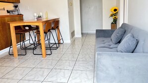 A living room with a grey sofa, a wooden dining table with chairs, a kitchen area with wooden cabinets, and a door leading to another room.