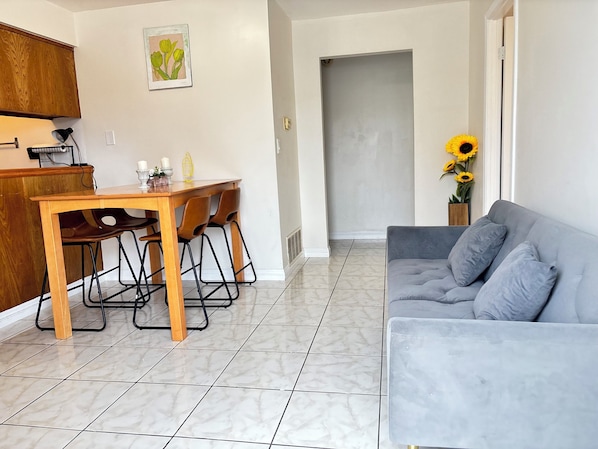 A living room with a grey sofa, a wooden dining table with chairs, a kitchen area with wooden cabinets, and a door leading to another room.