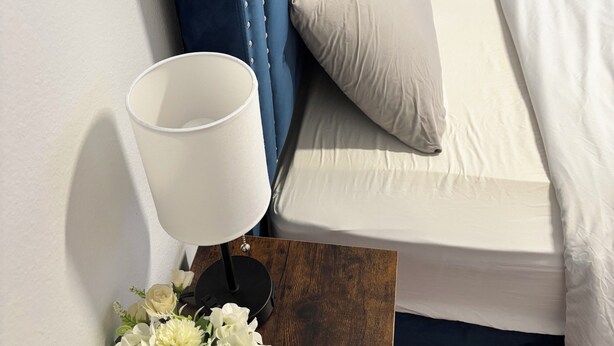A bed with a blue headboard, a white pillow, and a white blanket. There is a white lamp on a wooden nightstand with a flower arrangement beside it.