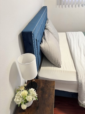 A bed with a blue headboard, a white pillow, and a white blanket. There is a white lamp on a wooden nightstand with a flower arrangement beside it.