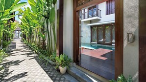 Ein Weg, gesäumt von großen Grünpflanzen, führt zu einer Holztür mit einem Balkon, der auf einen Pool blickt.