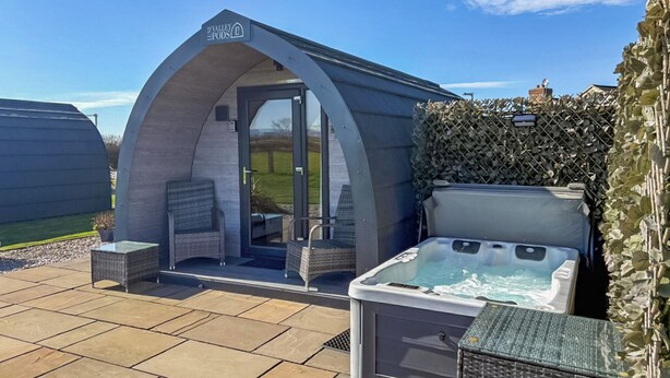 A hot tub and seating area with a covered entrance, surrounded by a patio with stone tiles and a hedge. - The Welbury - UKC9285 (Brompton)