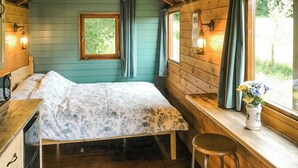 Une chambre de style cabane, avec un lit, des murs en bois, une fenêtre avec des rideaux, une petite table et un tabouret.