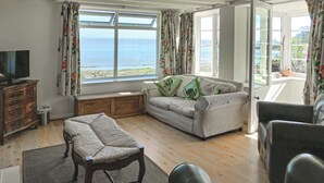 Ein Wohnzimmer mit Sofa, Fernseher, einer Holzbank und Meerblick durch große Fenster.