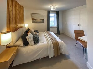 Habitació d'hotel moderna amb un llit gran, una taula de treball, una cadira, una làmpada, un quadre i una finestra amb cortines.
