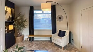A modern living room with a wooden herringbone floor, a white armchair with a dark cushion, a small wooden table, a potted plant, and a large window with blinds. - 2 bedroom accommodation in Newcastle Upon Tyne (Newcastle Upon Tyne)