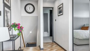 Une chambre d’hôtel moderne avec un parquet en bois disposé en motif chevrons, un grand lit avec une literie blanche, une table de chevet avec une lampe et une horloge murale.