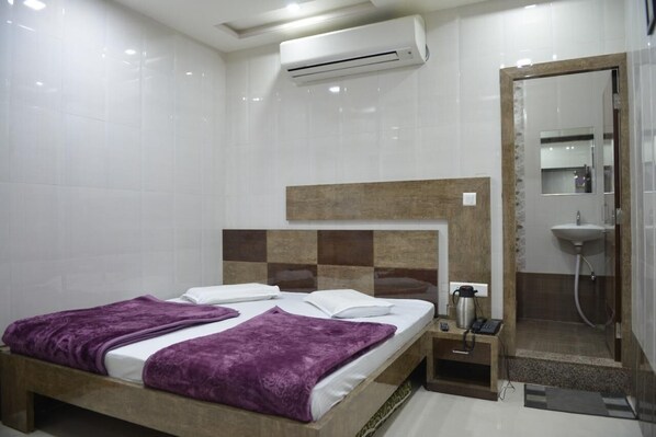 A hotel room with a bed, a bedside table with a phone and a coffee maker, an air conditioner, and a shower area.