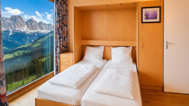 Un letto matrimoniale con testiera in legno, un comodino, un quadro incorniciato e una finestra con vista sulle montagne.