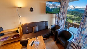 Un soggiorno moderno con divano, poltrone, tavolino, televisore, orologio e un balcone vista montagna.