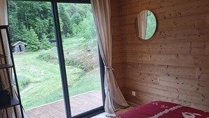 A bedroom with a bed, a wooden ceiling, a window with curtains, a circular mirror, and a view of a garden. - Chalet des Briseux (Saint-Maurice-sur-Moselle)