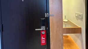 Quarto de hotel com uma porta escura, uma placa vermelha de 'NÃO ENTRE' e uma estante de madeira com um vaso.