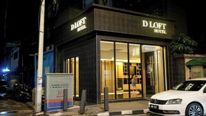 Hotel D Loft com fachada moderna, grande entrada de vidro e um carro branco estacionado na frente.