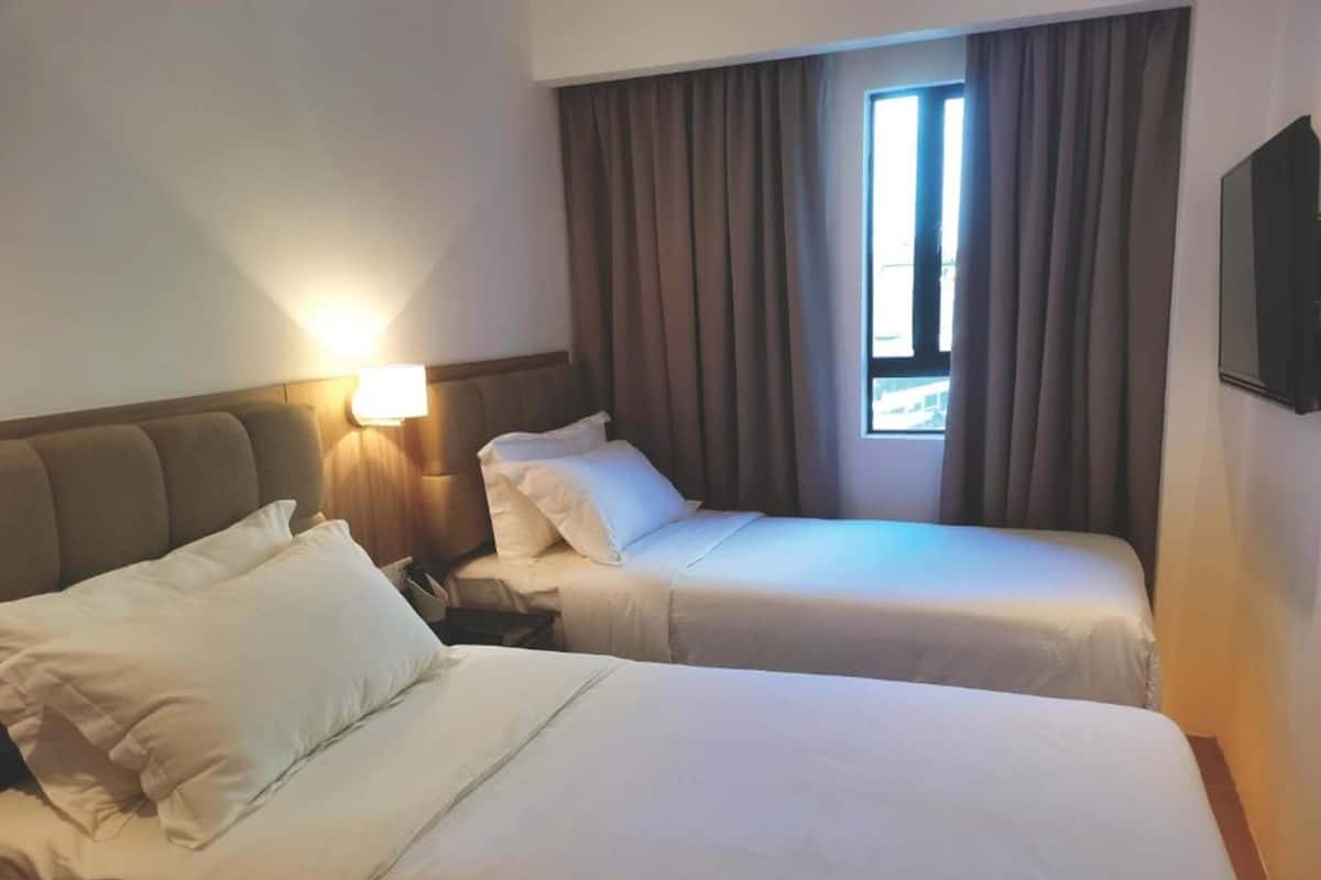 Une chambre d’hôtel avec deux lits, une table de chevet, une lampe, une fenêtre avec des rideaux et un téléviseur fixé au mur.