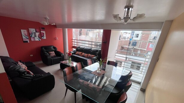 A modern living room with a glass dining table, black leather sofas, and red walls.