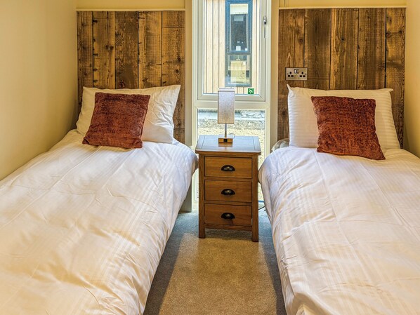 A room with two beds, wooden headboards, a nightstand with a lamp, and a window with a view.
