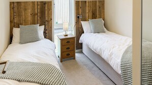 A bedroom with two beds, wooden headboards, a bedside table with a lamp, and a view through a window. - 3 bedroom accommodation in Perranporth (Perranporth)
