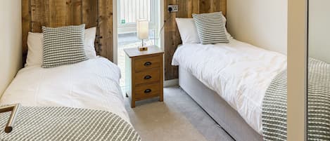 A bedroom with two beds, wooden headboards, a bedside table with a lamp, and a view through a window.